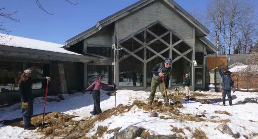 a group of students holding garden tools work on a piece of land in front of a building during a service project with outward bound